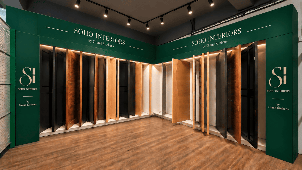An interior showroom corner with dark green branded walls displaying multiple wood-look door or panel samples in black, brown, and warm wood tones. The floor is wood-look laminate, and the ceiling has track lighting.
